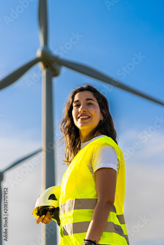 Female engineer holding a hard hat, smiling, standing in front of wind turbines