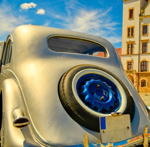 Classic silver sedan with a spare tire mounted on the back, parked near a historic building. A vintage automobile gleams under a bright blue sky with fluffy clouds, showcasing its iconic design and