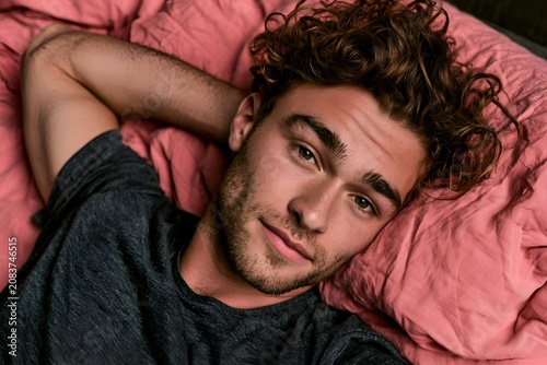 Relaxed handsome young man with curly hair lying on a pink bed looking at the camera