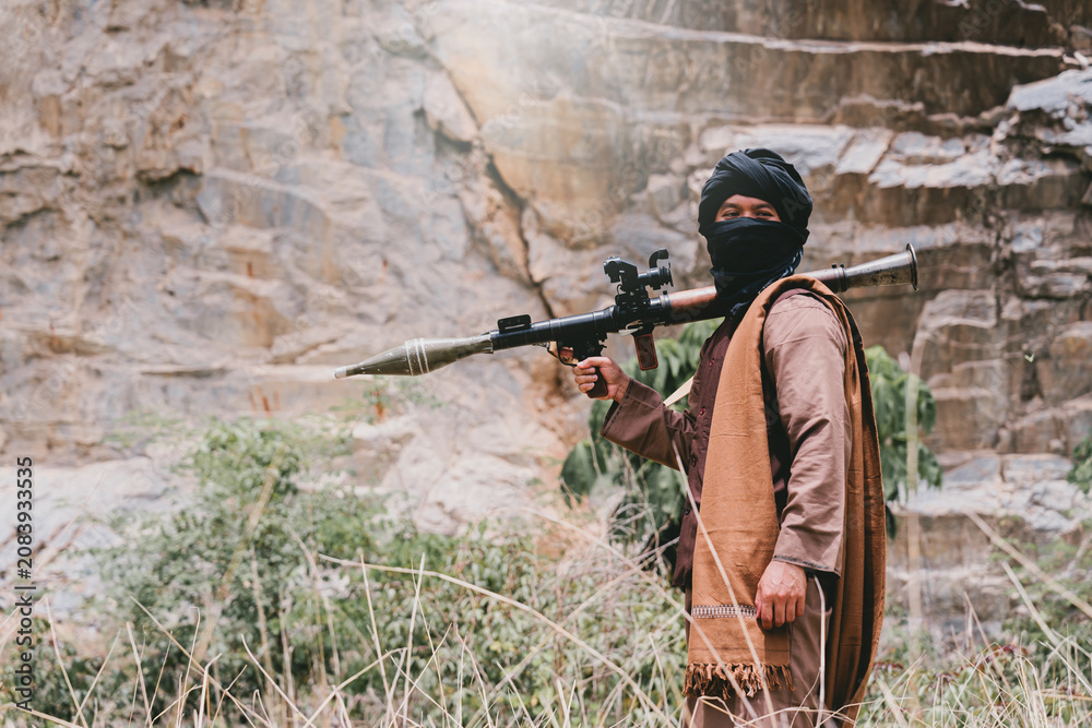 Fototapeta premium Outdoor military concept. Armed man in rocky canyon holding rocket launcher, masked face and desert clothing