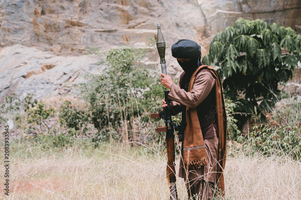 Fototapeta premium Outdoor military concept. Armed man in desert field with rifle and rocket launcher, masked warrior standing alert