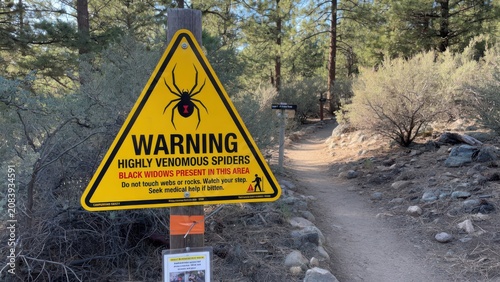 A yellow triangular danger sign stands on a wooden post along a mountain trail to warn hikers about the presence of venomous black widow arachnids that could be lurking in the nearby rocky