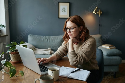 Young woman with glasses working on laptop at wooden table in cozy home living room with greenery.
