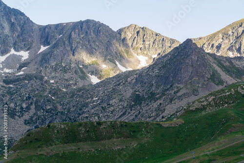 Jagged rocky Pyrenean peaks streaked with lingering snow rise above steep dark scree slopes in high mountains of Andorra. A vivid green alpine meadow sweeps across the foreground beneath a clear sky