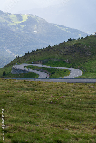 A serpentine mountain road carves dramatic S-curves through lush green alpine meadows rising into the Pyrenean peaks. Layered ridges and hazy summits frame breathtaking high-altitude road landscapes