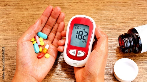 Top view of hands holding a digital glucometer device showing a high blood sugar level reading of 182 mg/dL alongside various colorful medical capsules over a wooden surface.