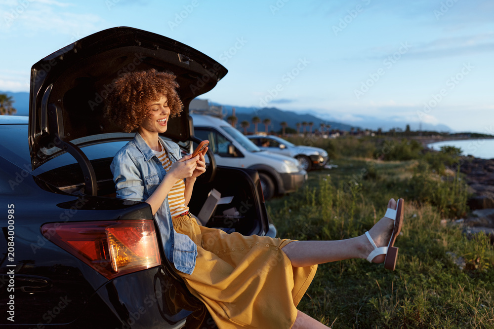 Fototapeta premium Woman with curly hair smiles while sitting on a car trunk and using a smartphone in an outdoor sunset scene, highlighting lifestyle travel outdoor sunset, casual relaxation and warm mood. Soft