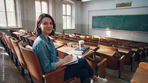 Young female student sitting in empty wooden lecture hall with books and coffee smiling at camera during study break.