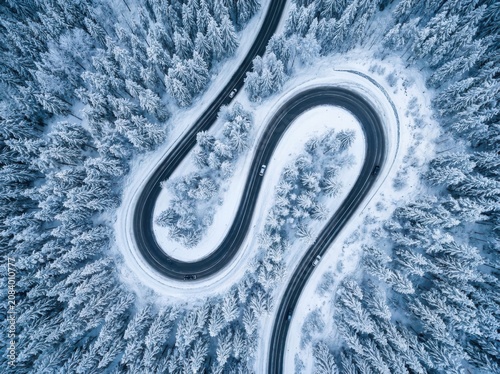 Curving winter road winding through snow covered pine forest from above with vehicles driving