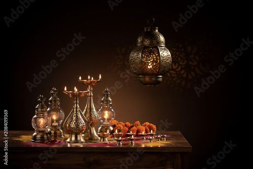 A group of traditional Indian brass oil lamps and ornate metal lanterns illuminate a festive display of vibrant orange marigold flowers and scattered colorful powders on a rich dark wooden surface