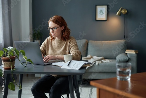 Young woman wearing glasses typing on laptop at desk while drinking coffee in comfortable home office during sunny morning.