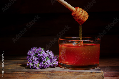Honey slowly dripping from a wooden dipper into a glass, next to wild thyme purple flowers on a rustic wooden surface