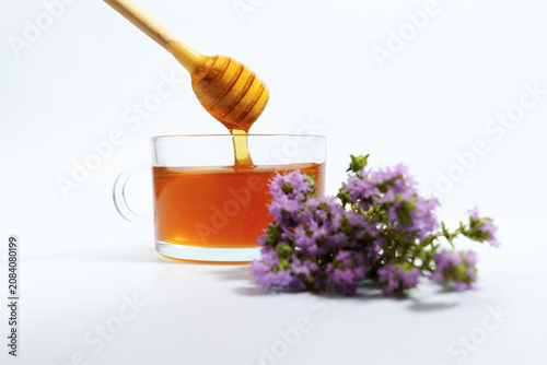 Honey dipper dripping sweet, natural honey into a glass cup of herbal infusion with fresh purple thyme flowers
