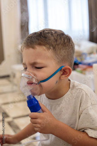 Young boy in white t-shirt using blue nebulizer inhaler with transparent mask and teal strap at home, steam visible during inhalation treatment for respiratory illness, cough, allergy or virus