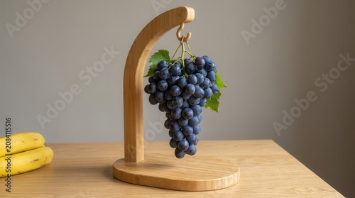 Cluster of dark blue grapes hanging from a wooden fruit holder on a light wooden table with bananas visible in the background