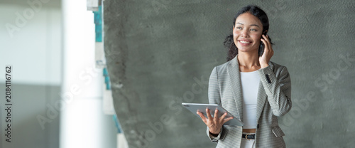 Portrait of smiling young businesswoman talking on mobile phone while holding digital tablet for business communication in modern office environment professional corporate people lifestyle.