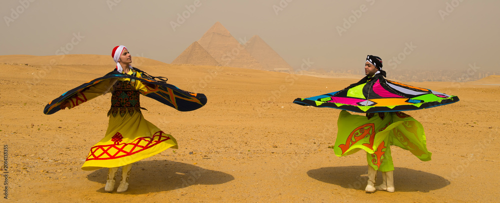 Fototapeta premium Two tanoura whirling dervish performers in vivid yellow and neon costumes spinning in exuberant cultural dance before the ancient Pyramids of Giza