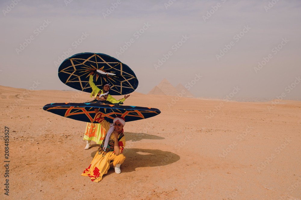 Fototapeta premium Two tanoura dance performers in vivid embroidered costumes fan spinning skirts dramatically before pyramid silhouettes in open Egyptian desert ideal