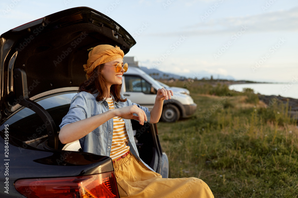 Naklejka premium Cheerful woman outdoors, sitting at a car trunk, sunlit moment, casual dress and scarf, carefree lifestyle, breezy afternoon, scenic coastline background