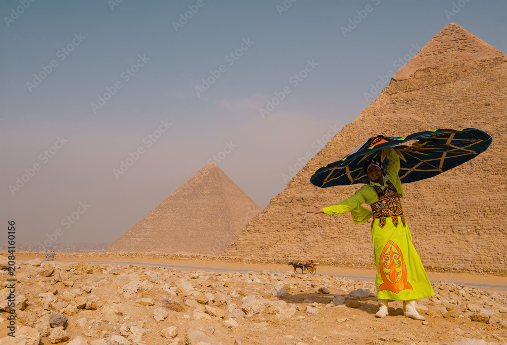 Fototapeta premium Festive egyptian folk performer in neon yellow embroidered costume balances large decorative umbrella before the iconic ancient pyramids on the rocky
