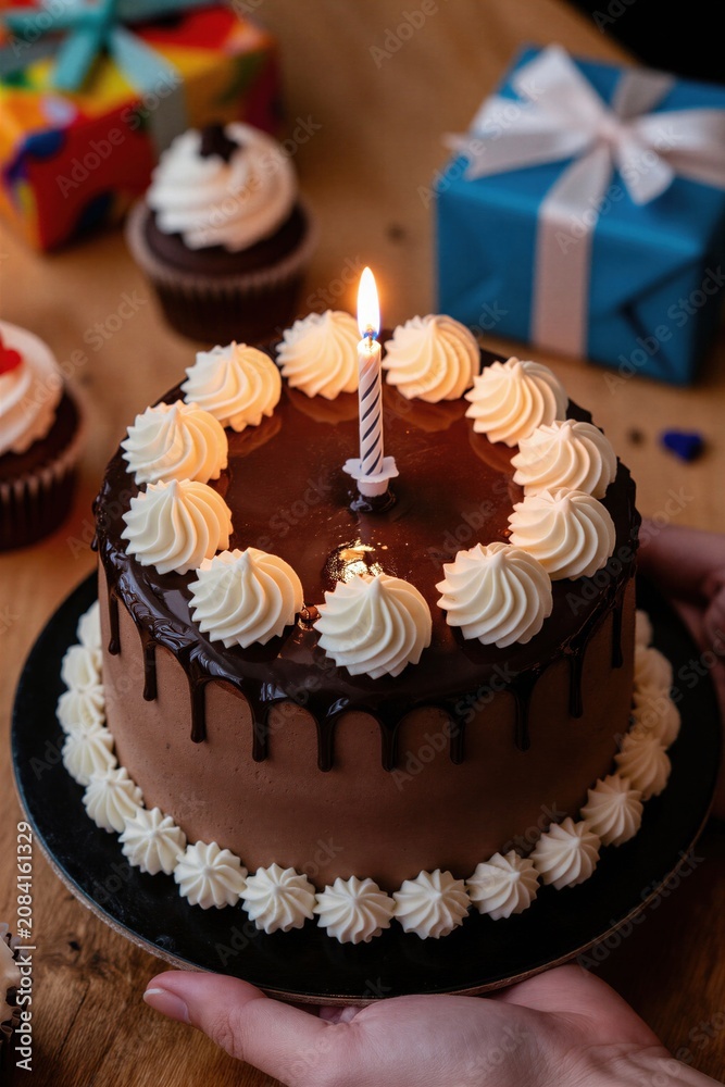 Fototapeta premium Chocolate birthday cake with single lit candle awaits wishes. Cupcakes and wrapped gifts surround dessert on wood table. Joyful intimate celebration scene.