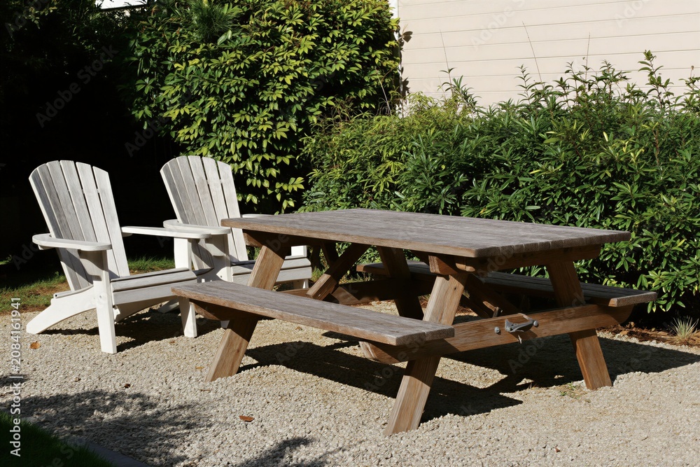 Fototapeta premium Wooden picnic table with benches, Adirondack chairs sits on gravel path. Rich green bushes, light colored building wall form background. Outdoor seating area offers relaxation, natural setting.