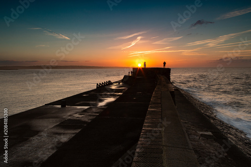 Obraz na plátne Photographers on the pier at Sunrise, silhouettes against a burgeoning sun, wispy orange clouds, and a turbulent sea