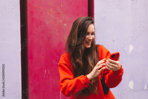 Happy laughing young woman in profile view looking at smartphone screen typing message, joyful female influencer using mobile phone app or watching funny online content near bright magenta wall