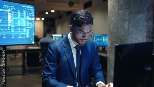 Young male broker wearing glasses and blue suit typing on keyboard at desk. Trader watching financial charts on monitors in dark office. Businessman working with stock market data late at night.