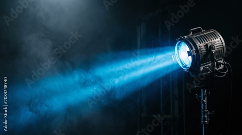 Fototapeta Studio lighting equipment projecting intense blue beam light through dark hazy atmosphere film production cinematic stage effect dramatic spotlighting photography backdrop visual performance.