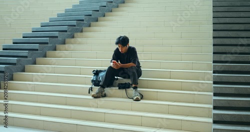 Young man waiting on wide staircase while checking smartphone, seated beside backpack on empty steps in minimal interior with repeating stair pattern and clean architectural lines.