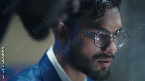 Close up of handsome businessman in glasses and looking at computer screen. Male trader analyzing financial data in dark office. Focused broker working with stock market and listening to colleague.