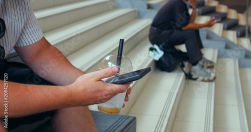 Hands using smartphone for mobile payment while holding plastic cup drink on indoor stairway, with seated people checking phones in background and modern public space details visible nearby