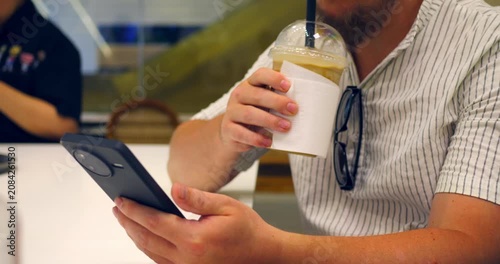 Man using smartphone while holding cold coffee drink in cafe, checking order on screen with takeaway cup and plastic lid, casual customer at table with eyeglasses hanging from shirt.