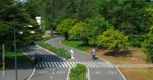 Cyclists ride along winding park path with zebra crossing and lane markings, surrounded by lush green trees, shrubs, and sunny outdoor recreation setting, showing active leisure travel through park.