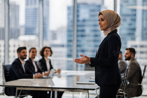 Successful Turkish muslim businesswoman in a hijab giving a corporate presentation to a diverse executive team in a modern glass boardroom