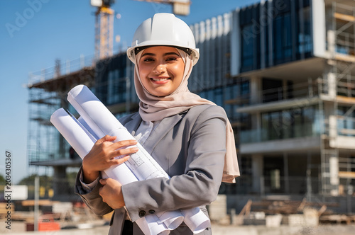 Confident female muslim architect wearing a hardhat and hijab holding blueprints while smiling at a modern construction site