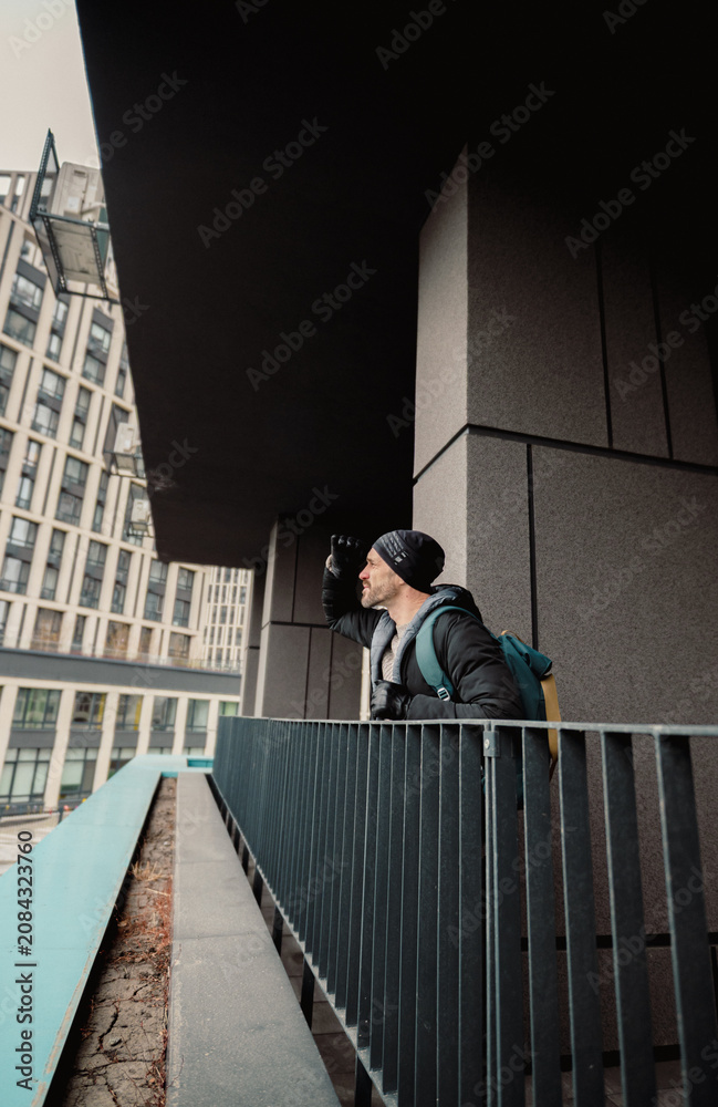 Fototapeta premium Modern Urban Explorer: Man with Backpack Looking Out from City Balcony on a Cloudy Day