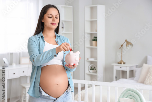 Young pregnant woman putting coin into piggy bank in childrens bedroom. Mater...