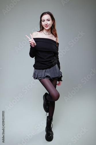 Studio fashion: smiling young woman dressed in skirt, jersey, boots and tights against gray background. Portrait of gorgeous merry girl showing 