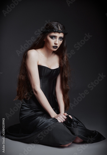 Studio portrait of the beautiful sad goth girl sitting on her knees. Lovely informal (gothic) lady dressed in black corset and skirt, with crown of thorns on head