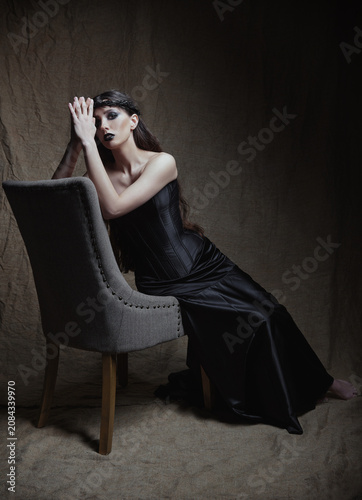 Adorable sad goth girl sitting on chair against the sackcloth background. Gorgeous informal (gothic) lady dressed in black corset and skirt, with crown of thorns on head