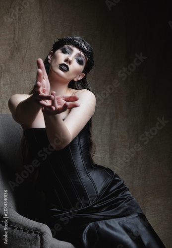 Beautiful sad goth girl sitting on armchair against the sacking background. Cute informal (gothic) lady dressed in black corset and skirt, with crown of thorns on head