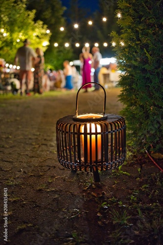 Decorative bamboo lantern with burning candle on the ground at a summer night garden party with blurred people background.