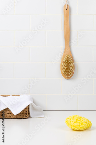 Minimalist bathroom mockup with a long handle wooden body brush hanging on a white subway tile wall. Yellow sea sponge and a white cotton towel in a wicker basket on the counter.