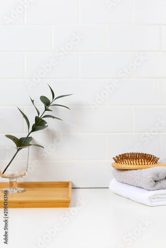 Minimalist bathroom product mockup with white subway tile background. Wooden tray with green leaf in glass and stacked towels with hairbrush for cosmetic display.