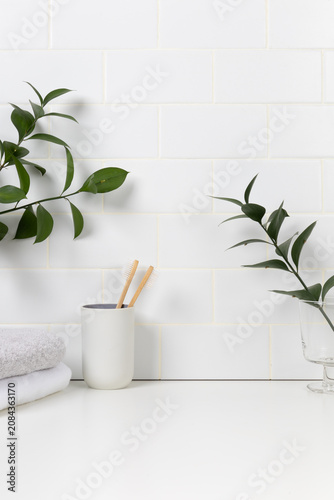 Eco-friendly dental care mockup background with white subway tile wall. Bamboo toothbrushes in a ceramic cup surrounded by fresh green leaves and soft folded towels.