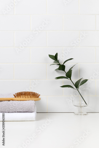 Minimalist bathroom product mockup with white subway tile background. Wooden tray with green leaf in glass and stacked towels with hairbrush for cosmetic display.