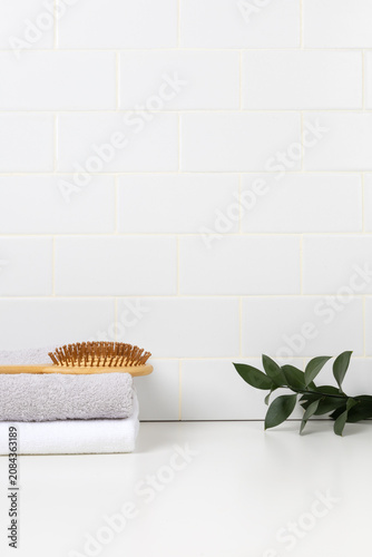Minimalist bathroom product mockup with white subway tile background. Wooden tray with green leaf in glass and stacked towels with hairbrush for cosmetic display.