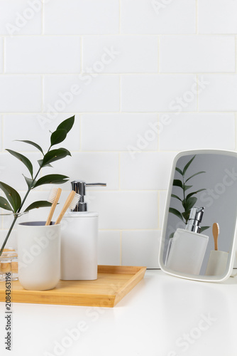 Eco-friendly oral care and bathroom amenities mockup. Bamboo toothbrushes, white soap dispenser on a wooden tray, and tabletop mirror with reflection against white subway tile wall.